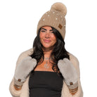 A woman wearing a beige knit beanie with a large pom-pom, pearl embellishments, and a small brown leather patch, paired with matching beige faux-fur gloves and a black top against a plain white background.