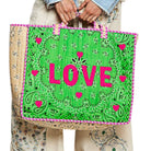 A person holds a large tote bag with a bright green paisley print, pink trim, and the word "LOVE" in bold pink letters across the front.