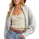 Woman wearing a beige tank top, gray cardigan, and blue jeans with a brown belt.