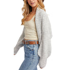 Woman wearing a light gray cardigan, beige tank top, blue jeans, and a brown belt on a white background