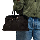 A person wearing an olive green jacket and blue jeans carries a dark brown suede shoulder bag with black faux-fur trim and gold-tone hardware against a white background.