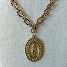 Gold chain necklace with an oval pendant featuring a religious figure surrounded by small clear stones, displayed on a textured off-white fabric background.