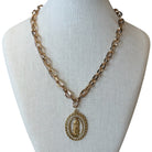 A gold-toned chain necklace with an oval pendant featuring a religious figure, displayed on a cream fabric mannequin bust against a white background.