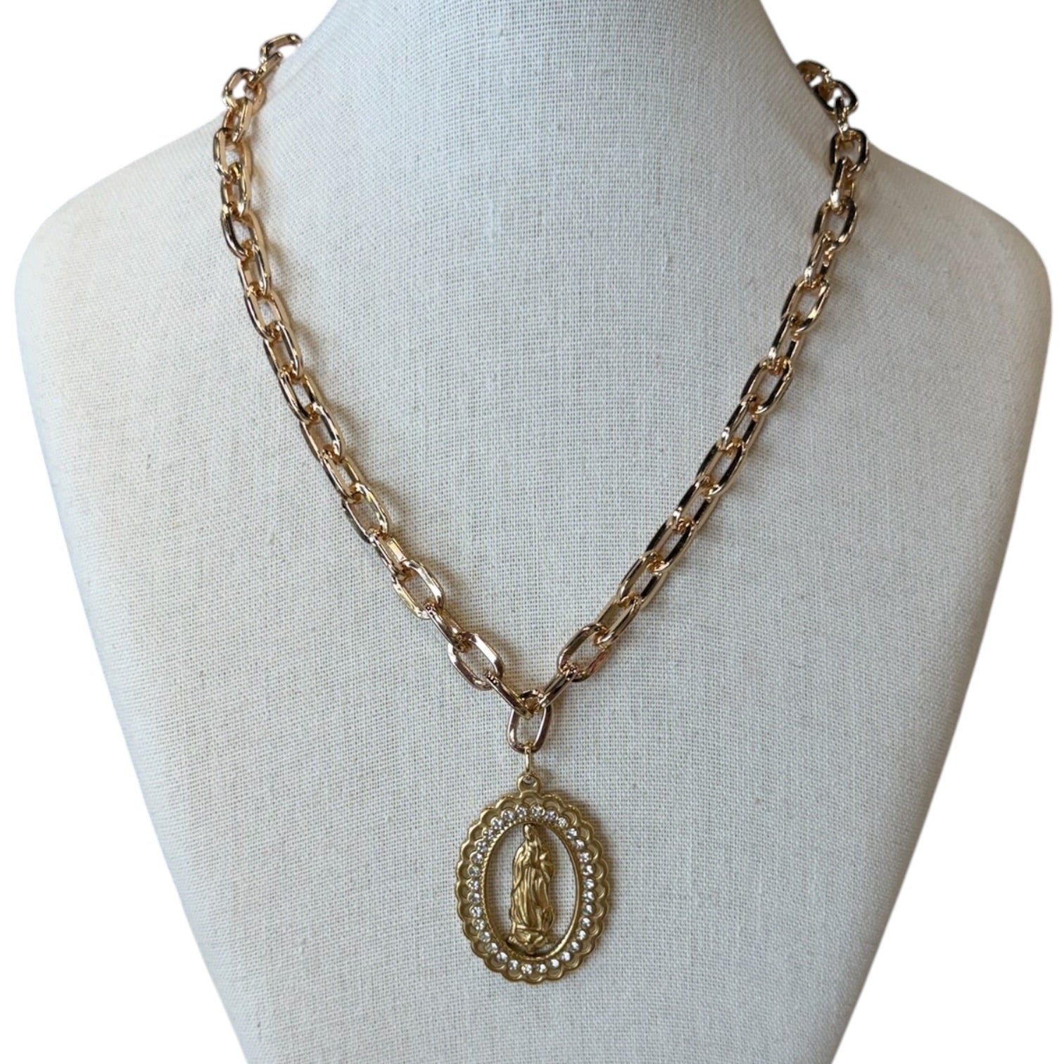 A gold-toned chain necklace with an oval pendant featuring a religious figure, displayed on a cream fabric mannequin bust against a white background.