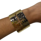A wide metallic cuff bracelet with a textured gold and black finish, featuring a cutout cross design on the front, shown worn on a person's wrist against a plain white background.