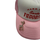 A close-up of a pink and white trucker hat featuring red embroidered text on the front panel and a rhinestone-embellished reindeer pin on the brim.