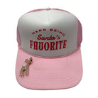 A pink and white trucker hat with red embroidered text reading 'HARD BEING Santa's FAVORITE' on the front panel, plus a small rhinestone reindeer pin on the brim, shown on a plain white background.