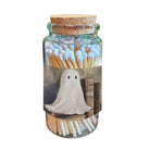 A clear glass jar with a cork stopper, filled with matchsticks and featuring a printed label of a white ghost standing in front of old books.