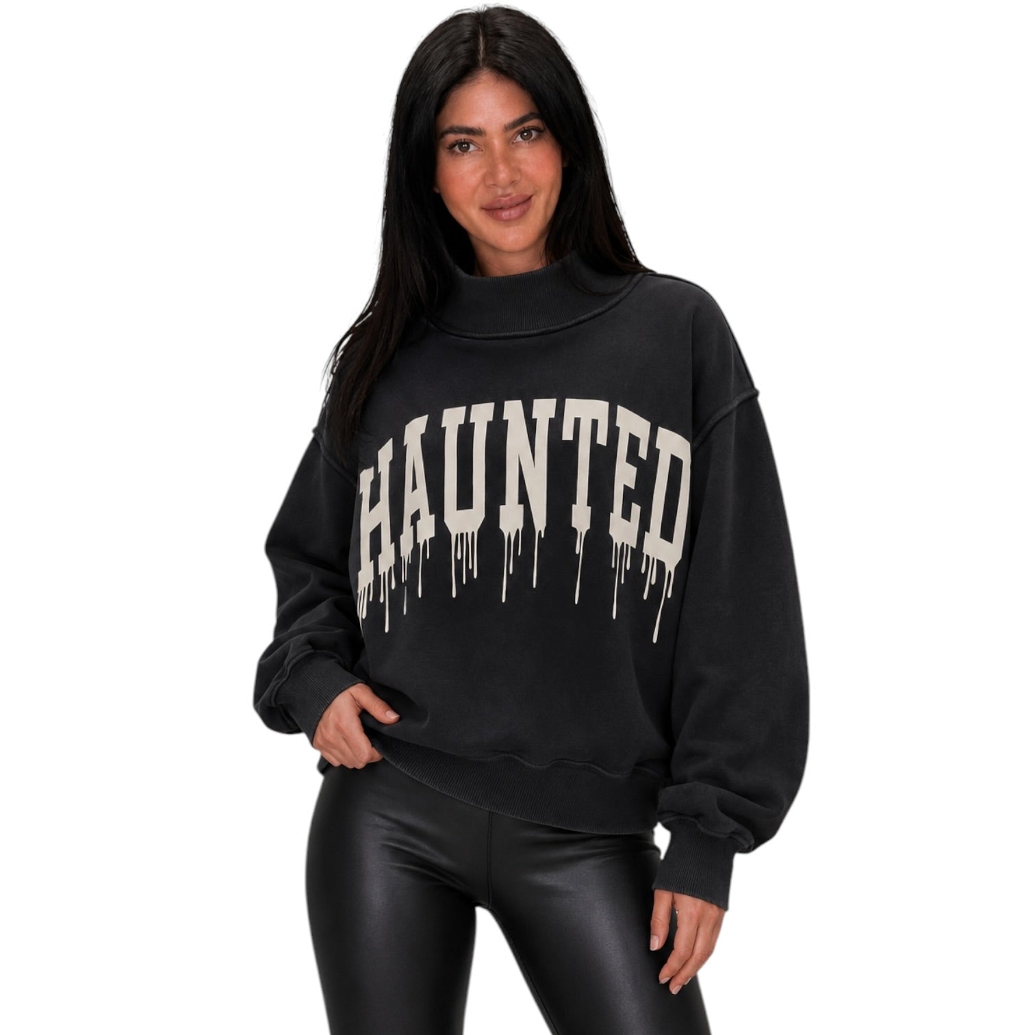 A woman modeling a black sweatshirt with a cream-colored 'HAUNTED' graphic printed across the chest in an arched, dripping font, paired with black faux-leather pants, against a solid white background.