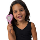 A young girl with dark hair smiling while holding a pink heart-shaped hairbrush with a glittery silver center, wearing a black sleeveless top against a plain white background.