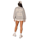 A woman shown from the back wearing a light gray knit varsity-style sweatshirt with cream lettering that reads 'Boston 1997 Everyday Moments.' She is also wearing a cream baseball cap, cream shorts, socks, and sneakers on a plain white studio background.
