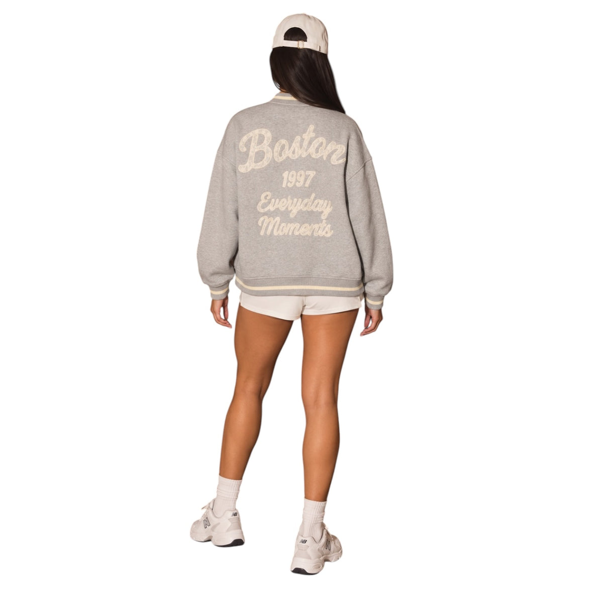 A woman shown from the back wearing a light gray knit varsity-style sweatshirt with cream lettering that reads 'Boston 1997 Everyday Moments.' She is also wearing a cream baseball cap, cream shorts, socks, and sneakers on a plain white studio background.