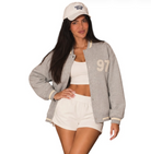 A woman modeling a light gray heather varsity jacket with cream ribbed trim and a cream '97' on the left chest, paired with a cream baseball cap, white crop top, and white shorts against a plain white background.