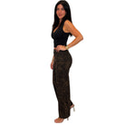 A woman modeling a pair of high-waisted, wide-leg pants with a brown and black leopard print pattern, paired with a black tank top and clear high-heeled sandals, set against a plain white background.