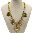 A gold-toned statement necklace displayed on a beige fabric mannequin bust. The necklace features a large Sacred Heart pendant, smaller heart charms, textured sunburst details, and small pearl-like bead accents.