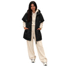 A woman modeling a black hooded puffer vest over a cream-colored hoodie and matching sweatpants, standing against a solid white background.