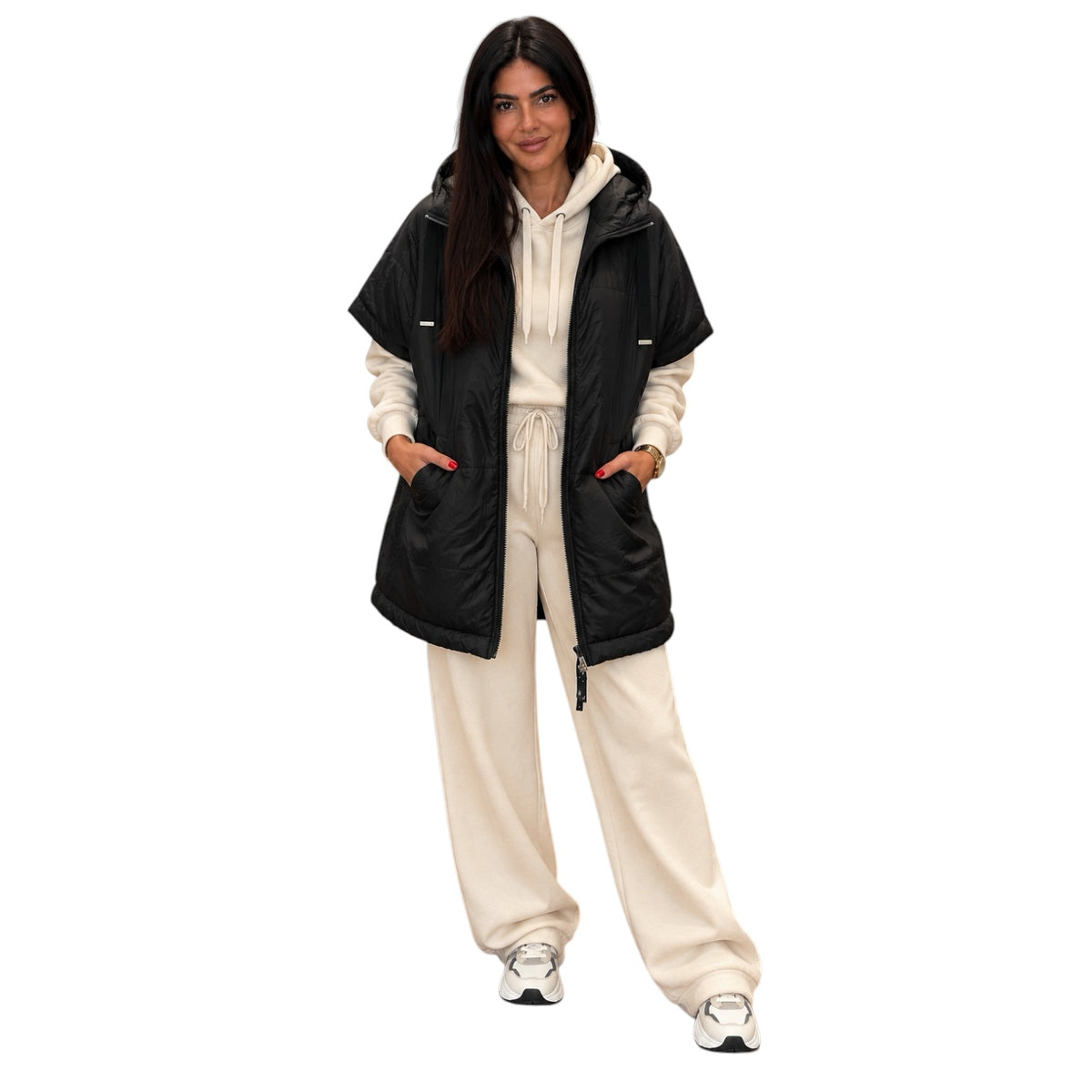 A woman modeling a black hooded puffer vest over a cream-colored hoodie and matching sweatpants, standing against a solid white background.