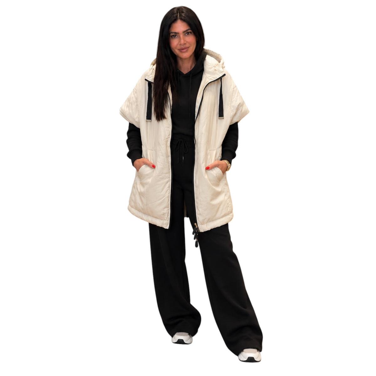 A woman modeling a cream-colored sleeveless puffer vest with a hood and front pockets, worn over a black outfit against a plain white background.