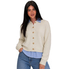 A woman modeling a cream-colored knit button-up cardigan sweater with brown buttons, layered over a light blue and white striped shirt and paired with blue jeans, against a solid white background.