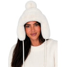 A woman wearing a fluffy off-white knit pom-pom beanie with ear flaps and drawstrings, paired with a matching cream knit sweater, against a plain white background.