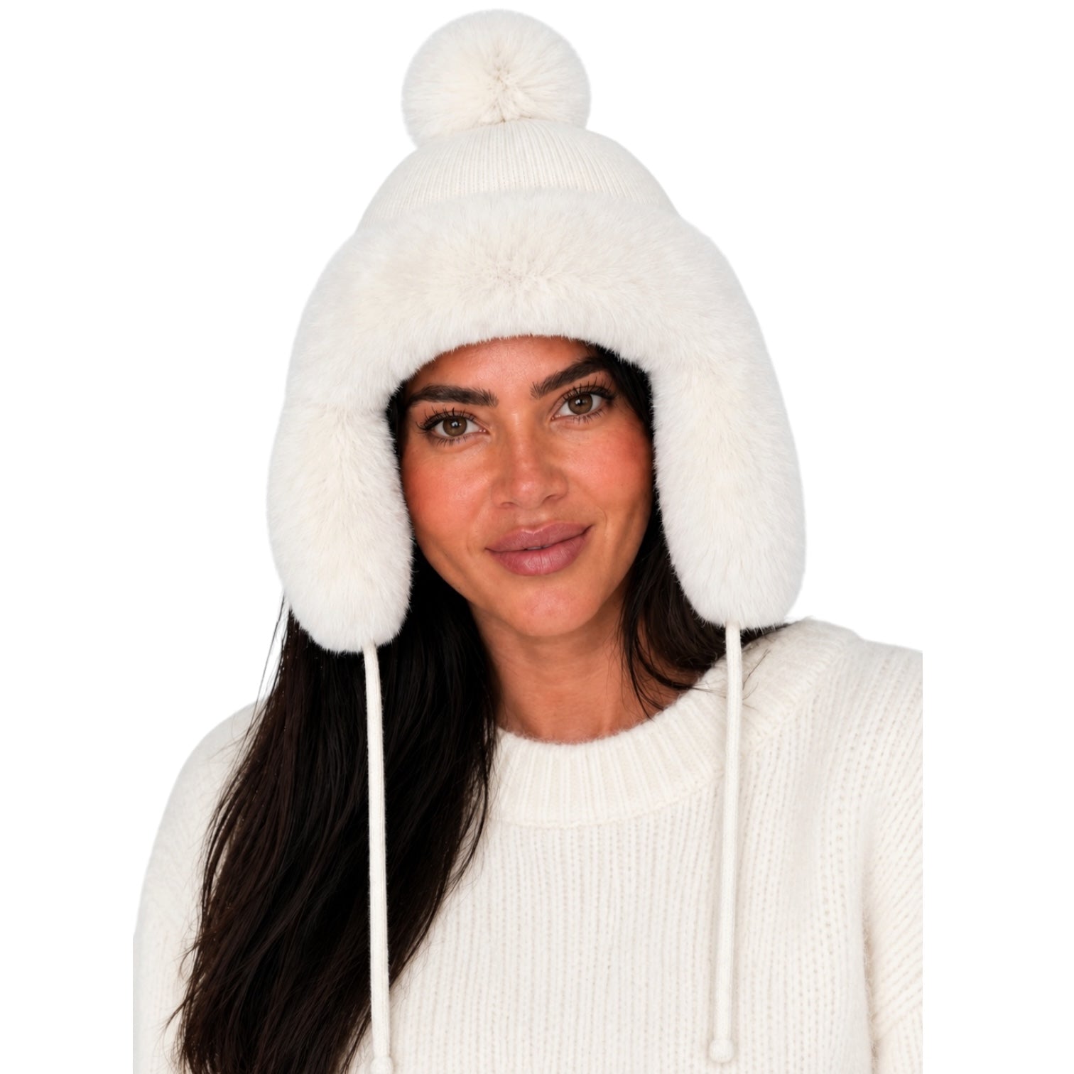 A woman wearing a fluffy off-white knit pom-pom beanie with ear flaps and drawstrings, paired with a matching cream knit sweater, against a plain white background.