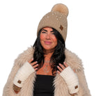 A woman wearing a beige knit beanie with a fluffy pom-pom and pearl-like embellishments, a cream faux-fur coat, and cream knit wrist warmers, shown against a plain white background.