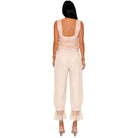 A woman viewed from the back wearing a sleeveless cream jumpsuit with lace trim at the back and ruffled lace hems, paired with clear high-heel sandals on a white background.