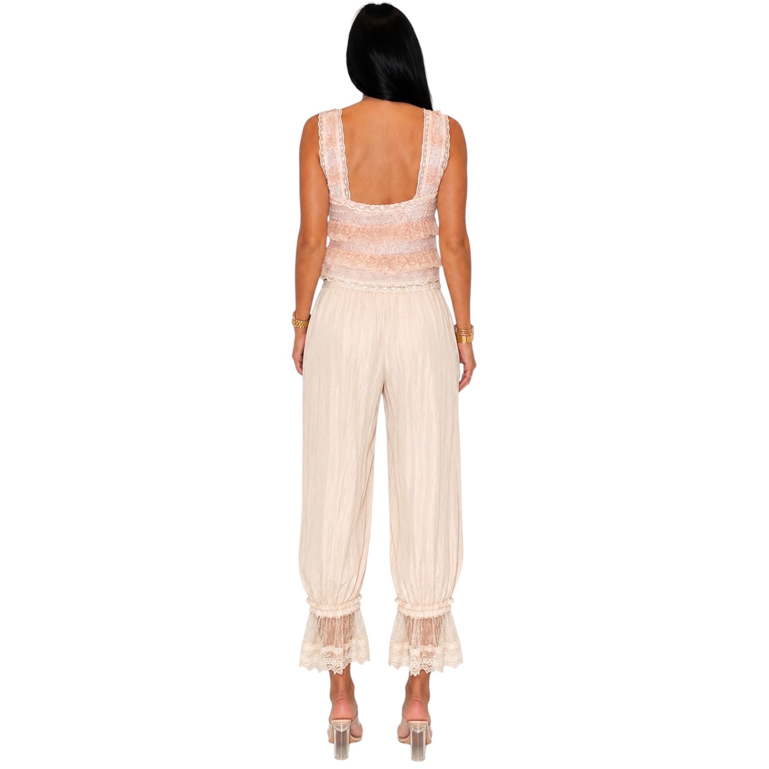 A woman viewed from the back wearing a sleeveless cream jumpsuit with lace trim at the back and ruffled lace hems, paired with clear high-heel sandals on a white background.