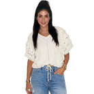 A woman modeling a cream-colored short-sleeve blouse with lace ruffle details and a drawstring tie at the waist, paired with light-wash blue jeans, against a plain white background.
