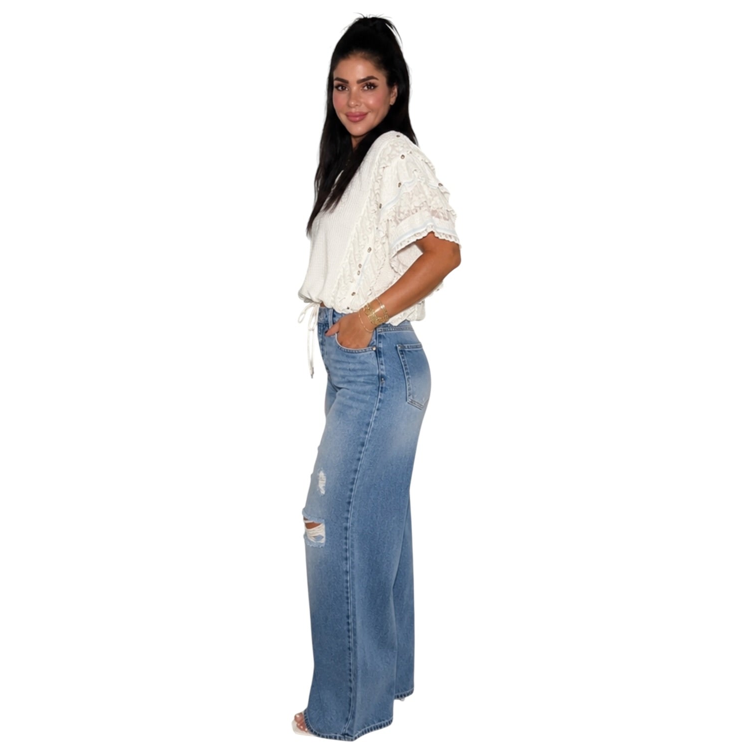 A woman modeling a white lace short-sleeve top and high-waisted, wide-leg blue denim jeans with distressed details, shown against a plain white background.