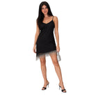 A woman modeling a black spaghetti-strap mini dress with a fringed hem, styled with black high heels and a small silver handbag against a plain white background.