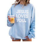 Person wearing a light blue sweatshirt with 'Jesus Loves You' text, holding a drink.