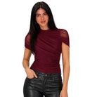 A woman modeling a ruched, short-sleeve top with sheer mesh shoulder panels in a deep burgundy color, paired with black faux-leather pants, against a solid white background.