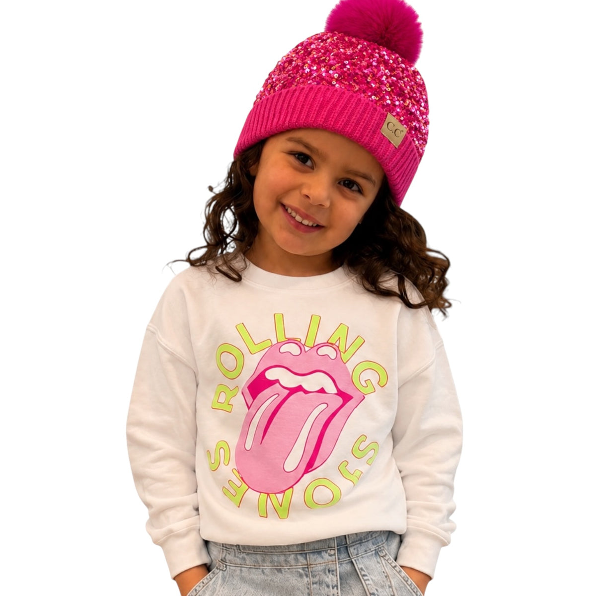 A young girl wearing a white long-sleeve sweatshirt with a pink and yellow Rolling Stones graphic, paired with a bright pink knit beanie featuring a pom-pom.