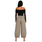 A woman shown from the back wearing a black off-the-shoulder long-sleeve top with lace sleeves and beige wide-leg pants with lace trim at the hems, against a plain white background.