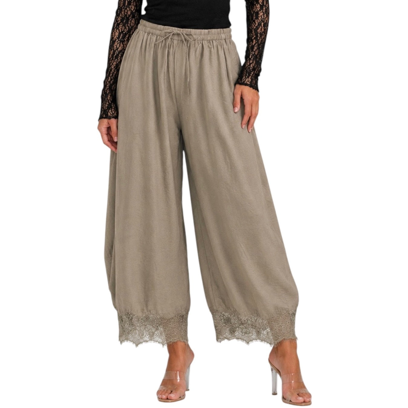A model wearing wide-leg beige pants with a lace hem, paired with a black lace long-sleeve top and clear high heels, shown against a plain white background.
