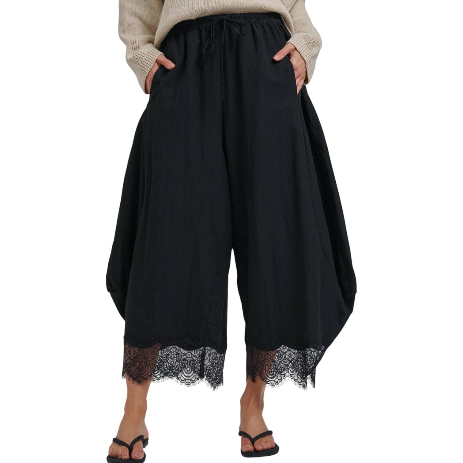 Black wide-leg cropped pants with a lace hem, modeled with a beige sweater and black flip-flops against a white background.
