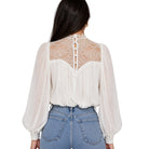 A woman viewed from the back wearing a white long-sleeve blouse with sheer puff sleeves, a lace back panel, and a buttoned lace neckline, paired with light blue jeans against a plain white background.