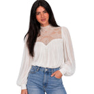 A woman modeling a white long-sleeve blouse with a sheer lace mock neck and puff sleeves, paired with blue jeans, against a plain white background.