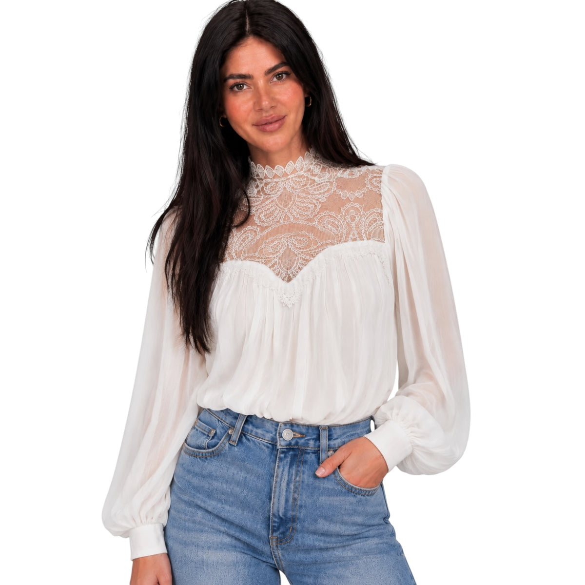 A woman modeling a white long-sleeve blouse with a sheer lace mock neck and puff sleeves, paired with blue jeans, against a plain white background.