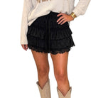 A person wearing a black ruffled lace mini skirt with a drawstring waist, paired with a cream sweater and beige cowboy boots, shown against a plain white background.