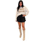 A woman modeling a cream sweatshirt with 'COFFEE WEATHER' printed in brown, paired with a black ruffled mini skirt and beige cowboy boots, against a solid white background.