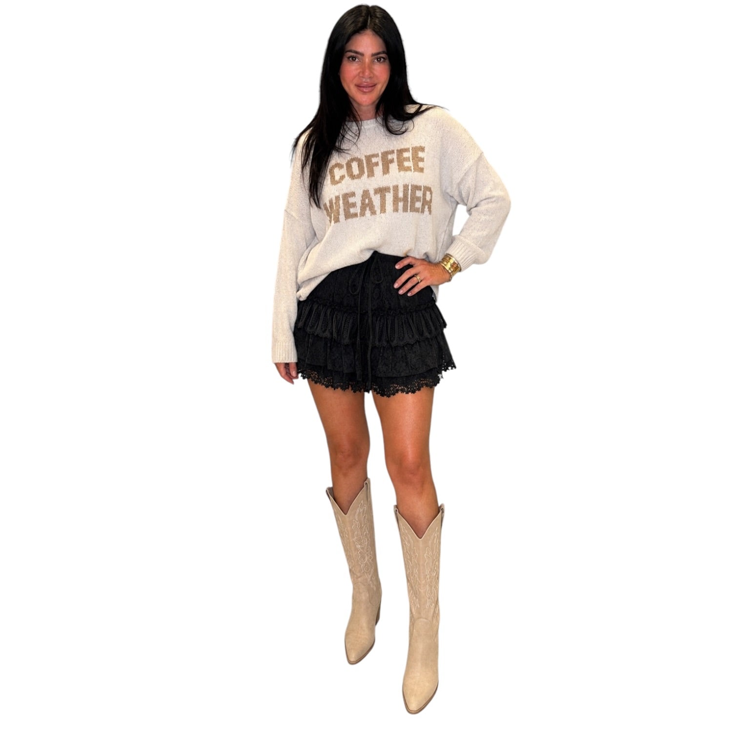 A woman modeling a cream sweatshirt with 'COFFEE WEATHER' printed in brown, paired with a black ruffled mini skirt and beige cowboy boots, against a solid white background.