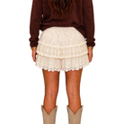 A person shown from the back wearing a cream lace tiered ruffle mini skirt, a dark brown long-sleeve top, and beige cowboy boots against a plain white background.