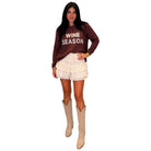 A woman modeling a dark brown sweatshirt with 'WINE SEASON' printed in white, paired with a cream lace tiered mini skirt and beige cowboy boots, against a solid white background.