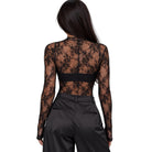 A woman shown from the back wearing a black sheer lace long-sleeve top with a black waistband, paired with black trousers, against a plain white background.