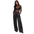 A woman modeling a black sheer lace long-sleeve crop top paired with high-waisted black satin wide-leg trousers, shown against a plain white background.