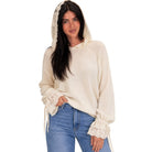 A woman wearing a cream-colored knit hoodie with lace trim on the hood and ruffled cuffs, paired with blue jeans, against a plain white background.