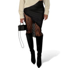 Person wearing a black lace skirt with a beige sweater and black knee-high boots on a white background