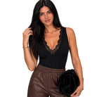 A woman modeling a black sleeveless bodysuit with a lace V-neckline, paired with brown pants and holding a black flower-shaped clutch against a plain white background.